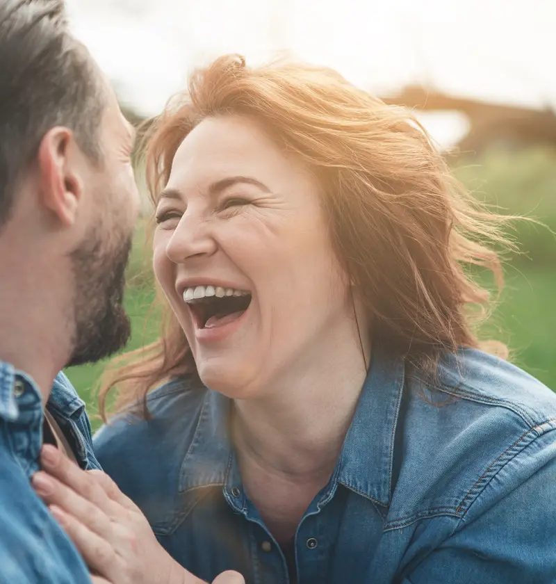 Cherishing Joyful Moments as a Carent – A middle-aged woman and man in denim jackets share a joyful moment outdoors, laughing together in a sunlit park. A theme of finding happiness and connection despite the challenges of caring for an elderly parent