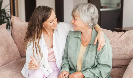 A smiling adult daughter sits close to her elderly mother on a sofa, sharing a warm moment and holding hands. They are looking at each other with affection, showing a strong, supportive relationship.