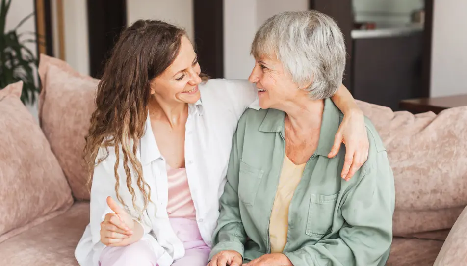 A smiling adult daughter sits close to her elderly mother on a sofa, sharing a warm moment and holding hands. They are looking at each other with affection, showing a strong, supportive relationship.