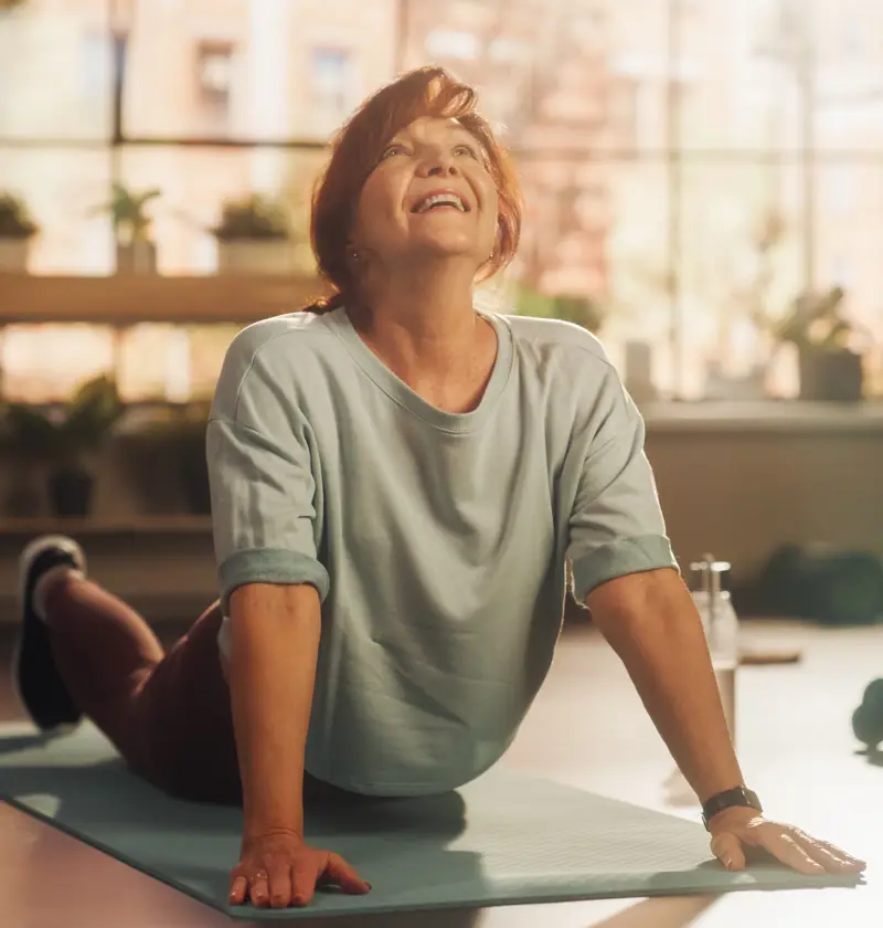 Self-Care and Wellbeing for Carents – A smiling woman in her 50s or 60s does a yoga stretch on a mat in a bright, cosy living room. A representation of self-care, stress relief, and maintaining wellbeing while caring for an elderly parent.
