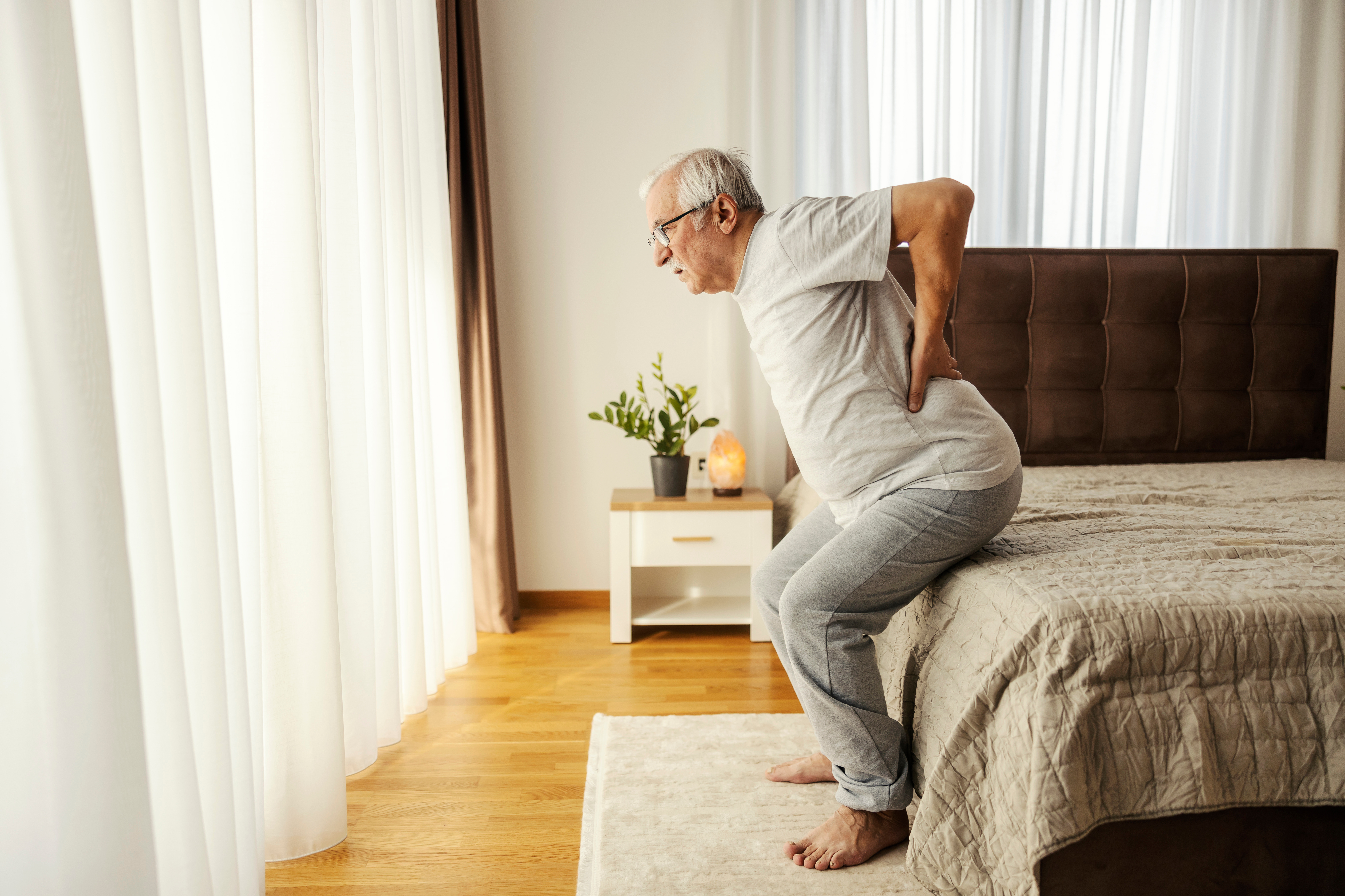 Older man sitting up from bed with visible back discomfort – highlighting the need for bed sensor pads to monitor movement, alert carers to changes, and support fall prevention for elderly parents.