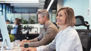 A smiling woman wearing a headset types on a keyboard in a bright office. A representation of working carers balancing employment with caring responsibilities and understanding workplace rights, flexible working, and carer support policies.