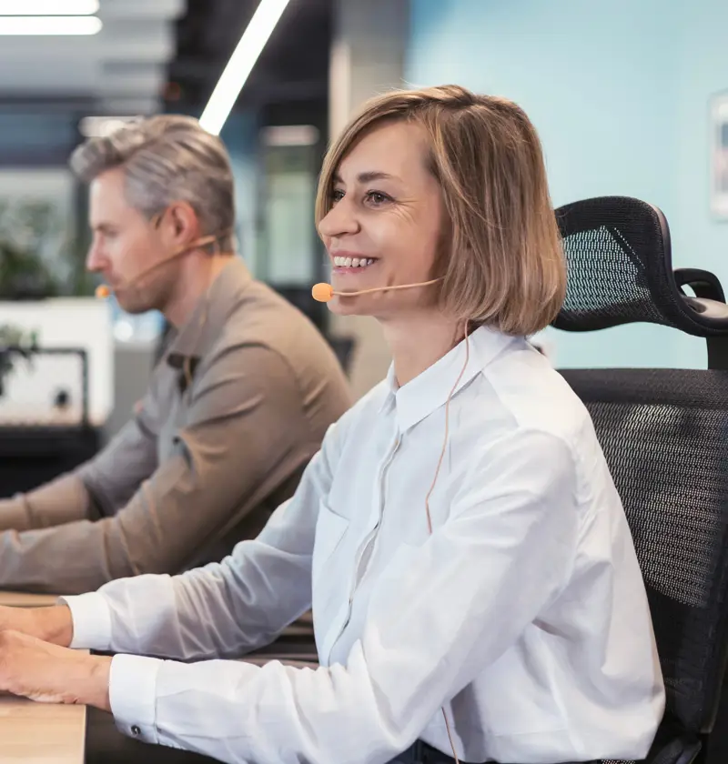 A smiling woman wearing a headset types on a keyboard in a bright office. A representation of working carers balancing employment with caring responsibilities and understanding workplace rights, flexible working, and carer support policies.