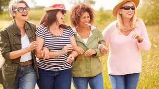 A group of four middle-aged women walking arm in arm through a countryside path, smiling and laughing. A reminder of the importance of friendship and emotional support for those caring for elderly parents.