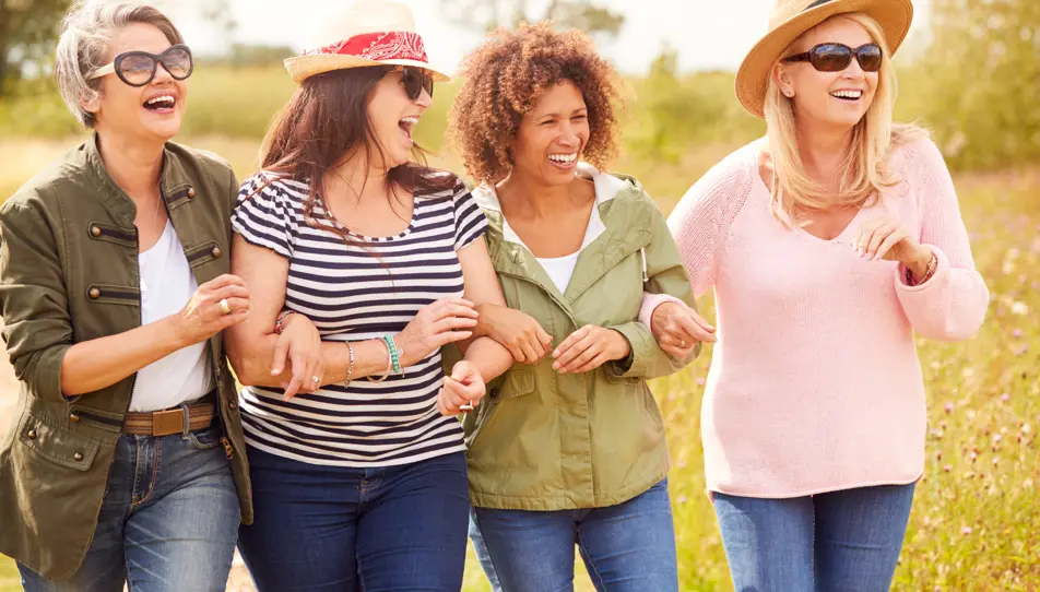 A group of four middle-aged women walking arm in arm through a countryside path, smiling and laughing. A reminder of the importance of friendship and emotional support for those caring for elderly parents.