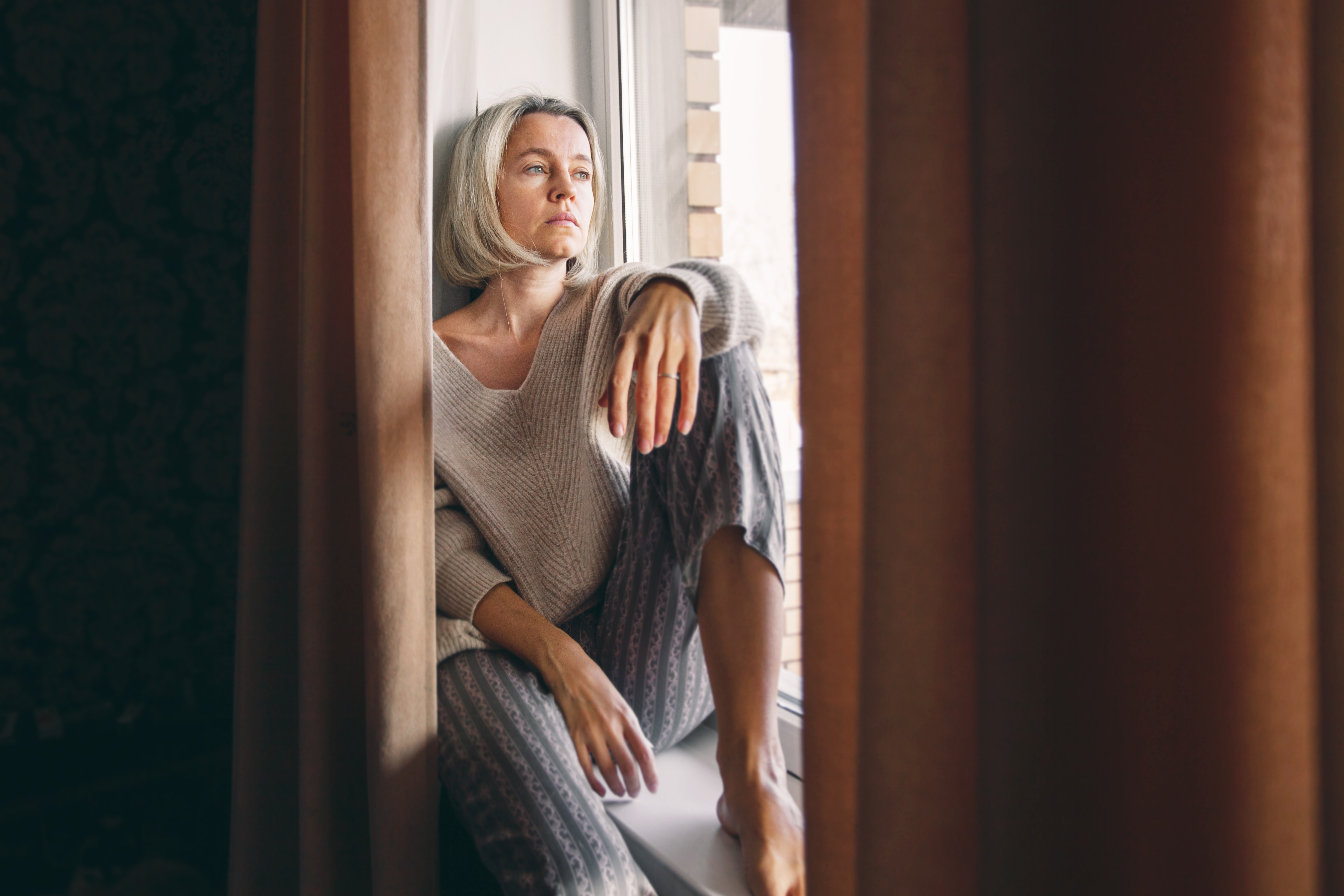 Reflecting on the Challenges of Caring for an eldery parent – A middle-aged woman in a warm sweater and patterned pyjama bottoms gazes out of a window with a pensive expression. A depiction of the isolation and quiet moments of reflection experienced by many caring for elderly parents.