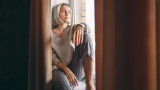 Reflecting on the Challenges of Caring for an eldery parent – A middle-aged woman in a warm sweater and patterned pyjama bottoms gazes out of a window with a pensive expression. A depiction of the isolation and quiet moments of reflection experienced by many caring for elderly parents.