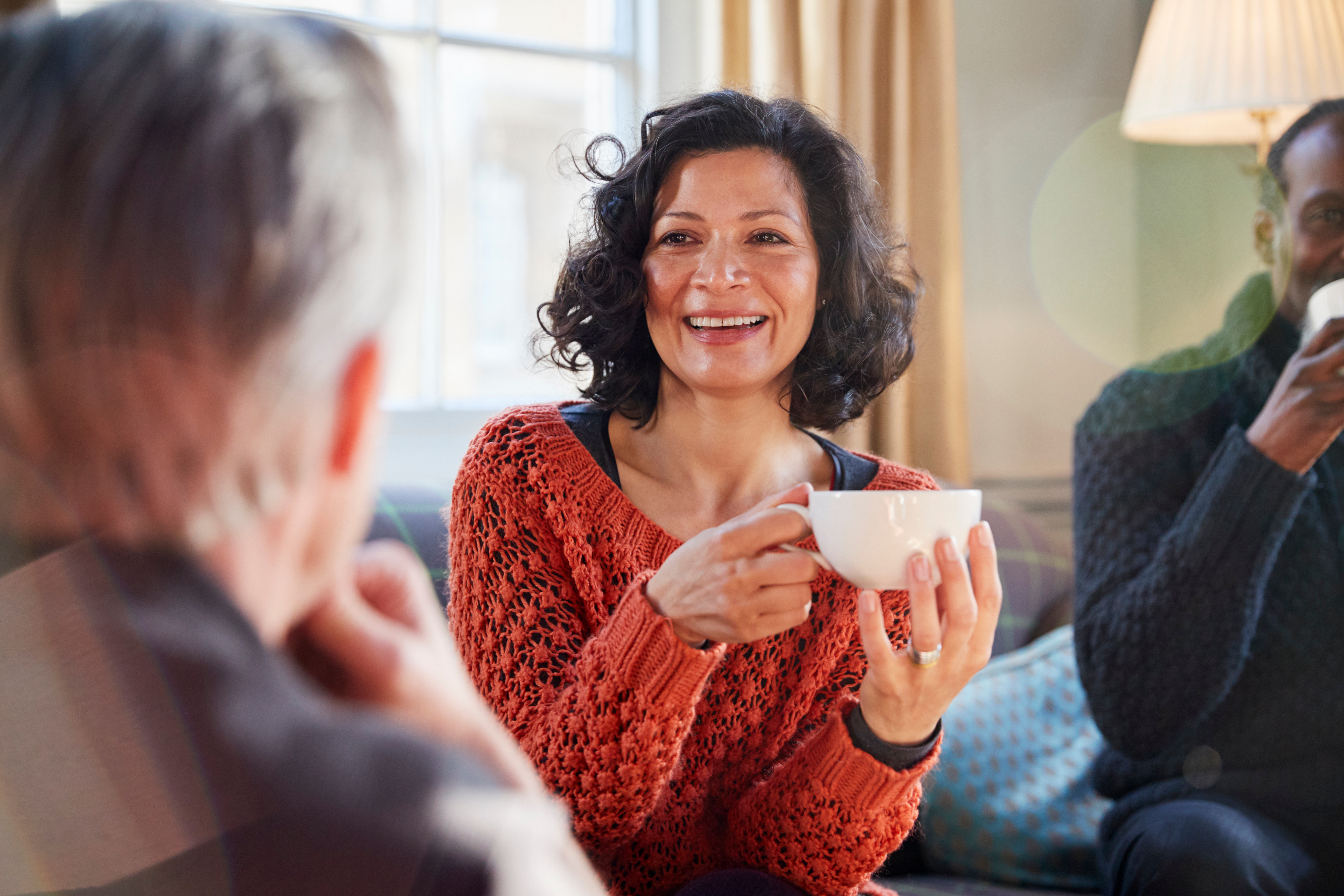 Support Networks: Carents Connecting Over Coffee – A cheerful woman in a burnt-orange knitted sweater holds a cup of coffee, engaging in a warm conversation with friends. A representation of emotional support, carer connections, and the importance of social interaction while navigating caregiving responsibilities.