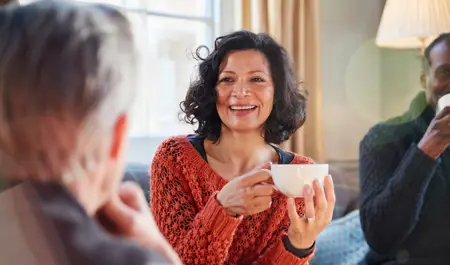 Support Networks: Carents Connecting Over Coffee – A cheerful woman in a burnt-orange knitted sweater holds a cup of coffee, engaging in a warm conversation with friends. A representation of emotional support, carer connections, and the importance of social interaction while navigating caregiving responsibilities.