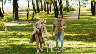 Taking a Break: Carents Enjoying Time Outdoors – A mature couple enjoys a peaceful walk in a lush green park, holding coffee cups while walking their golden retriever. A reminder of the importance of fresh air, companionship, and personal wellbeing for those supporting an ageing parent.
