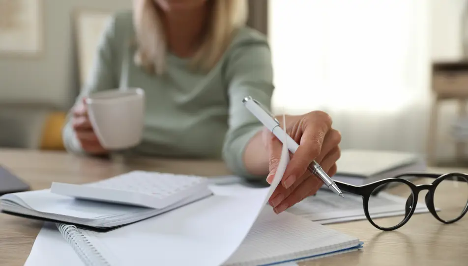 Managing Household Finances for Elderly Care – A woman reviews financial documents while holding a coffee cup, symbolising the responsibility of managing bills, care costs, and financial planning for ageing parents.