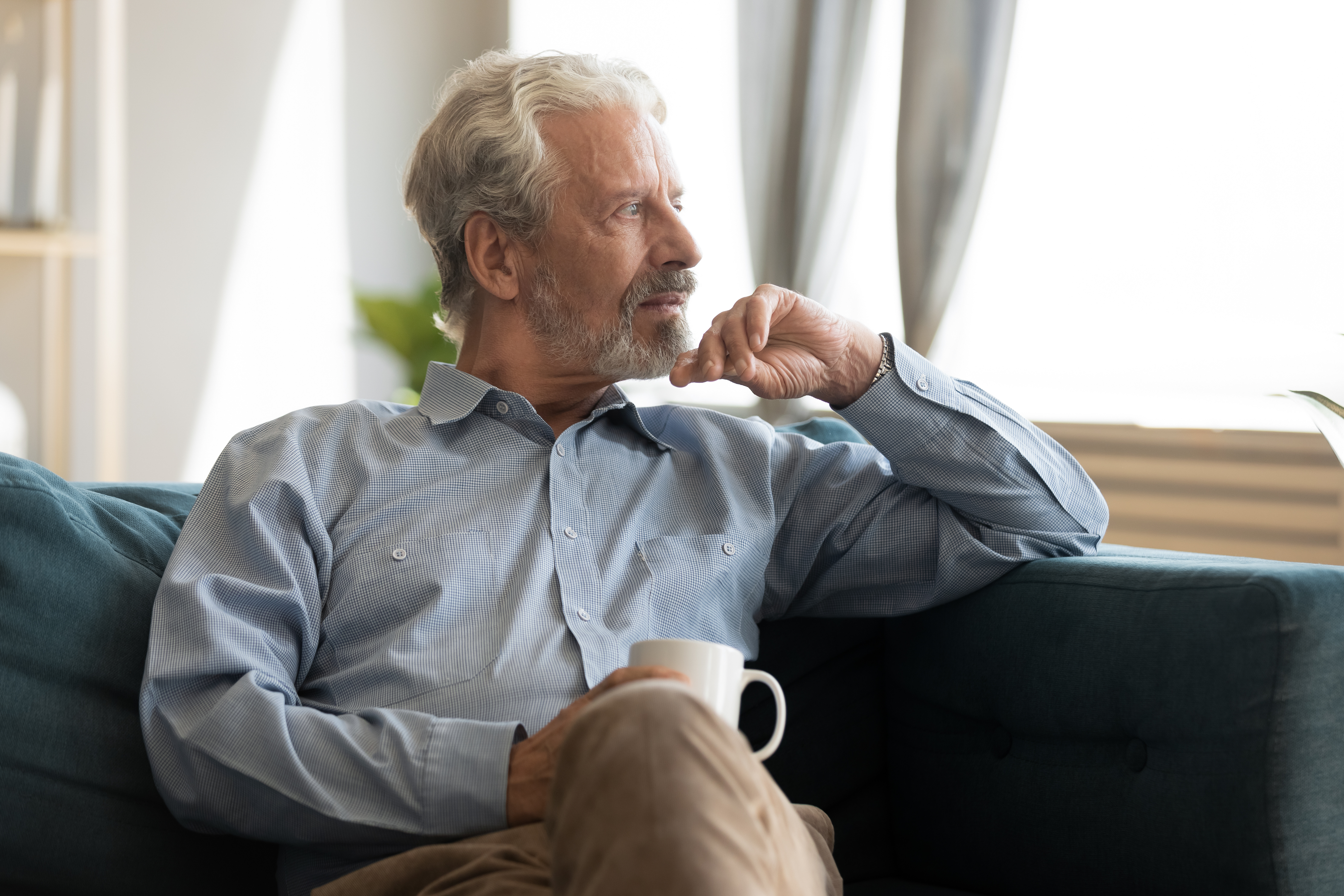 Finding Moments to reflect on your Emotions as a Carent – A silver-haired man sits on a plush sofa, holding a coffee cup while staring thoughtfully into the distance. A representation of taking time for oneself amidst the responsibilities of supporting an ageing parent.