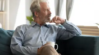 Finding Moments to reflect on your Emotions as a Carent – A silver-haired man sits on a plush sofa, holding a coffee cup while staring thoughtfully into the distance. A representation of taking time for oneself amidst the responsibilities of supporting an ageing parent.