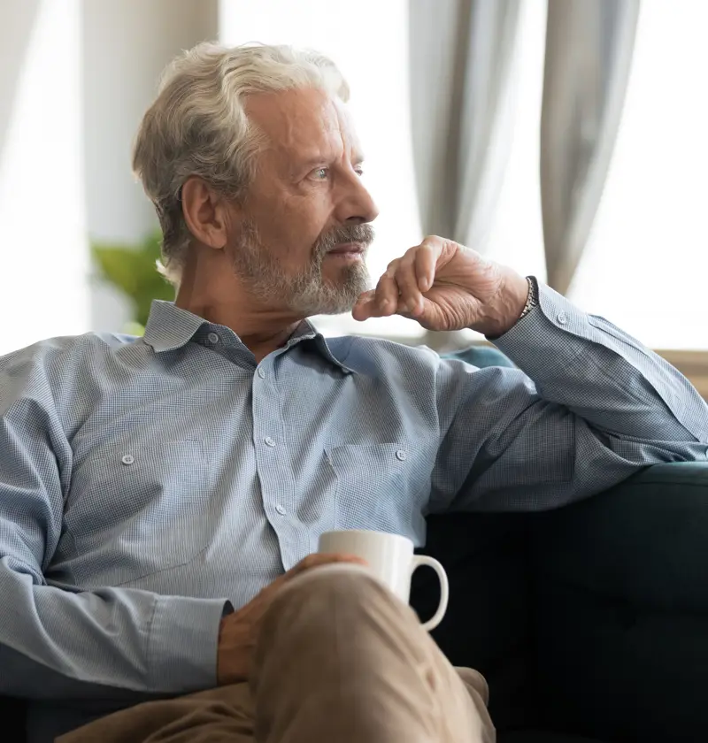 Finding Moments to reflect on your Emotions as a Carent – A silver-haired man sits on a plush sofa, holding a coffee cup while staring thoughtfully into the distance. A representation of taking time for oneself amidst the responsibilities of supporting an ageing parent.