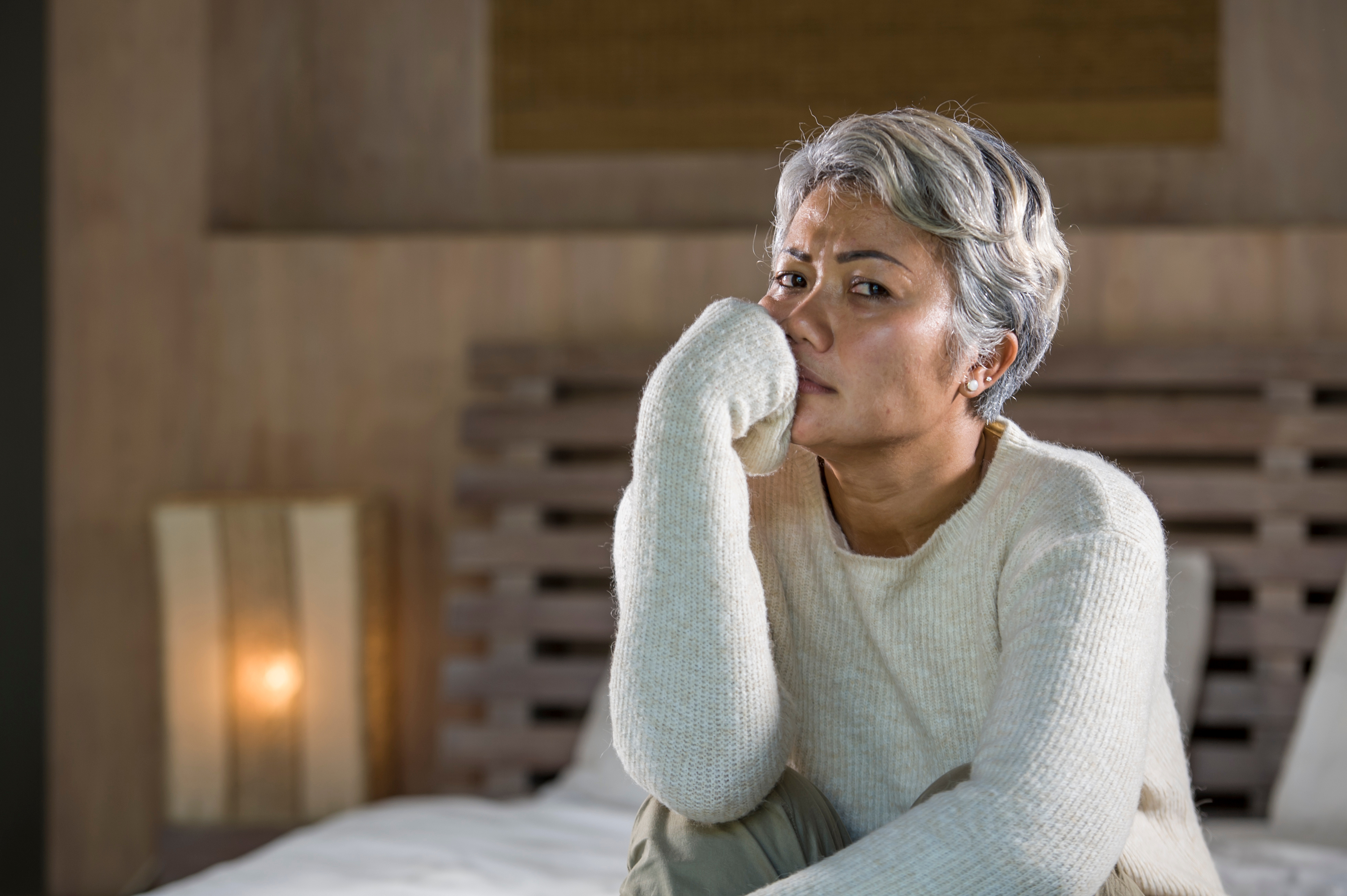 Navigating Stress and Change in Caregiving – A middle-aged woman with salt-and-pepper hair in a cream sweater sits on her bed, resting her hand on her mouth in concern. A scene highlighting the emotional complexities of caring for a parent and coping with change.