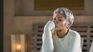 Navigating Stress and Change in Caregiving – A middle-aged woman with salt-and-pepper hair in a cream sweater sits on her bed, resting her hand on her mouth in concern. A scene highlighting the emotional complexities of caring for a parent and coping with change.