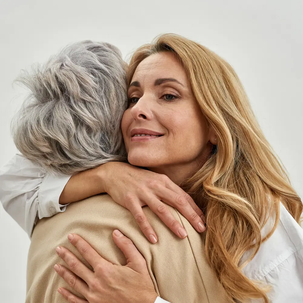 A woman embraces an elderly parent, both smiling gently as they share a moment of affection. The image conveys warmth, care, and emotional connection. The background is soft and neutral, focusing on the bond between them.