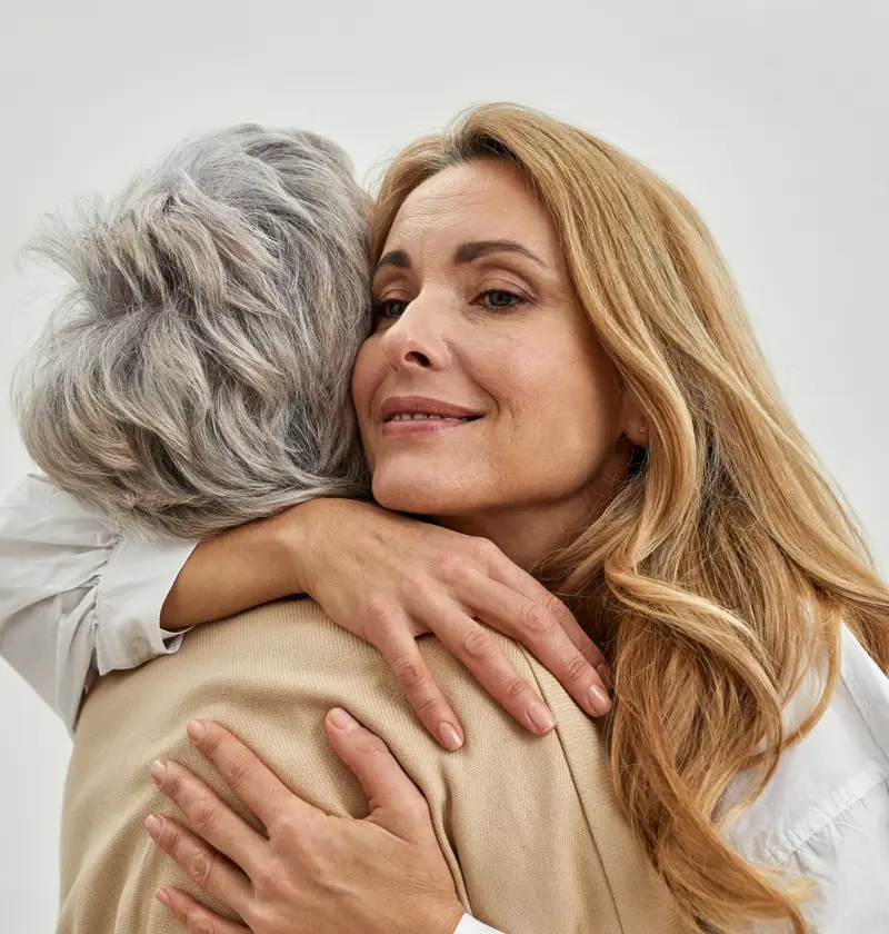 A woman embraces an elderly parent, both smiling gently as they share a moment of affection. The image conveys warmth, care, and emotional connection. The background is soft and neutral, focusing on the bond between them.
